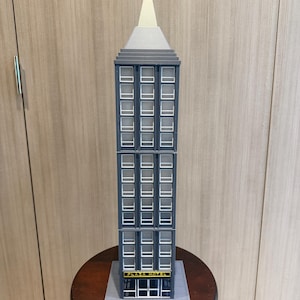 Può includere: Un modello in miniatura di un grattacielo, con una facciata grigio scuro e numerose finestre rettangolari. L'edificio è sormontato da una sezione grigio chiaro e da una guglia bianca appuntita. La base dell'edificio mostra il testo "PLAZA HOTEL" in giallo.