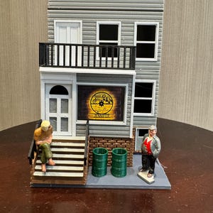 May include: A miniature gray house model with a black balcony, white doors and windows, and a sign that reads "NALO SALON JENNIFER". Two green barrels and two miniature figures are in front of the house.