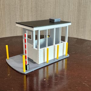 Puede incluir: Un modelo de caseta de peaje en miniatura con techo negro, paredes blancas y base gris. El modelo presenta postes de seguridad amarillos y rojos y una pequeña estructura gris en el techo. El diseño es una réplica detallada.