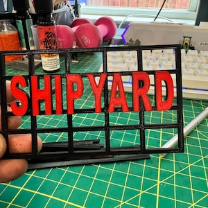 以下が含まれることがあります： 赤色の文字で「SHIPYARD」と書かれた黒色の3Dプリントサイン。サインは格子状の構造をしています。背景には、緑色のカッティングマット、キーボード、その他の事務用品があります。