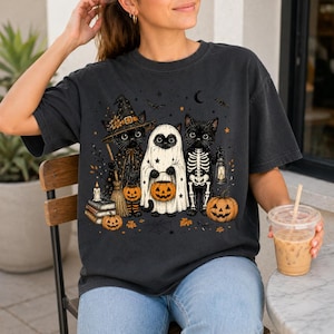 May include: A charcoal gray t-shirt featuring a graphic of three black cats dressed for Halloween. One cat wears a witch hat and holds a broom, one is dressed as a ghost holding a pumpkin bucket, and one wears a skeleton costume holding a lantern.