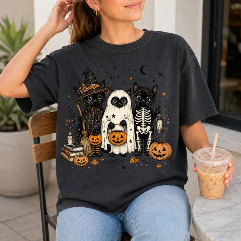 May include: A charcoal gray t-shirt featuring a graphic of three black cats dressed for Halloween. One cat wears a witch hat and holds a broom, one is dressed as a ghost holding a pumpkin bucket, and one wears a skeleton costume holding a lantern.