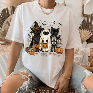 May include: A white t-shirt featuring a graphic of three black cats dressed for Halloween. One cat wears a witch hat and cloak, one is dressed as a ghost, and one wears a skeleton costume. They are surrounded by pumpkins, stars, and a crescent moon.