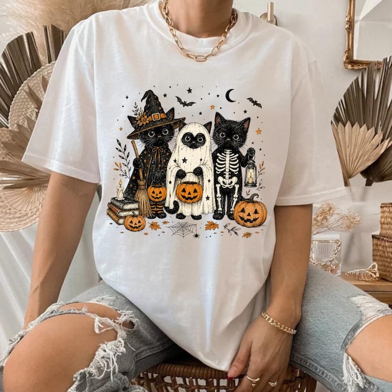 May include: A white t-shirt featuring a graphic of three black cats dressed for Halloween. One cat wears a witch hat and cloak, one is dressed as a ghost, and one wears a skeleton costume. They are surrounded by pumpkins, stars, and a crescent moon.