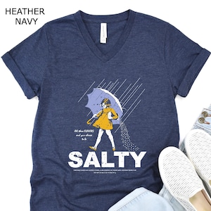 May include: A heather navy V-neck t-shirt featuring a graphic of a person in a yellow raincoat holding an umbrella in the rain, with the text "All the flavours and you choose to be SALTY" printed in white, paired with light blue jeans and white slip-on shoes.
