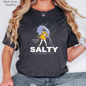 May include: A dark grey heather t-shirt featuring a graphic of a person in a yellow raincoat holding a blue umbrella under rain. The text reads All these FLAVORS and you choose to be SALTY, with additional small text below the word SALTY.
