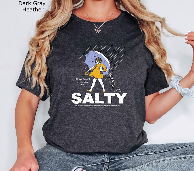 May include: A dark grey heather t-shirt featuring a graphic of a person in a yellow raincoat holding a blue umbrella under rain. The text reads All these FLAVORS and you choose to be SALTY, with additional small text below the word SALTY.
