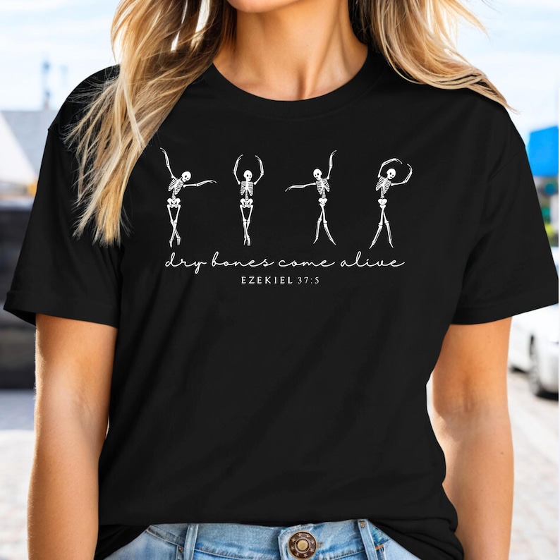 A black t-shirt with a graphic design featuring four white silhouettes of dancers performing a dance pose, accompanied by the text "dry bones come alive" and the Bible verse "EZKEL 1:3".