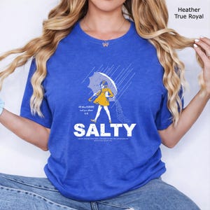 May include: A heather true royal blue t-shirt featuring a graphic of a person in a yellow raincoat holding an umbrella in the rain, with the text "All these FLAVORS and you choose to be SALTY" and additional small text printed below the graphic.