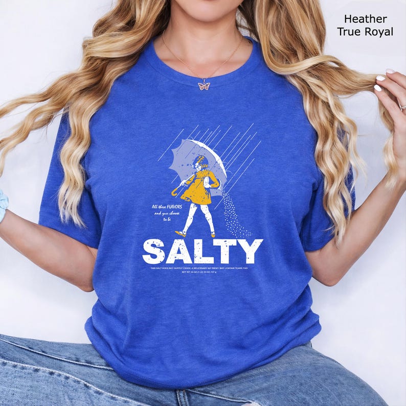 May include: A heather true royal blue t-shirt featuring a graphic of a person in a yellow raincoat holding an umbrella in the rain, with the text "All these FLAVORS and you choose to be SALTY" and additional small text printed below the graphic.