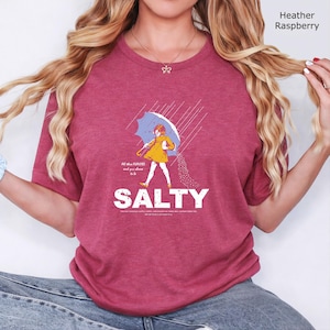 May include: A heather raspberry t-shirt featuring a graphic of a person in a yellow raincoat holding a blue umbrella in the rain, with the text "All these FLAVORS and you choose to be SALTY" and additional small text printed below the main word.