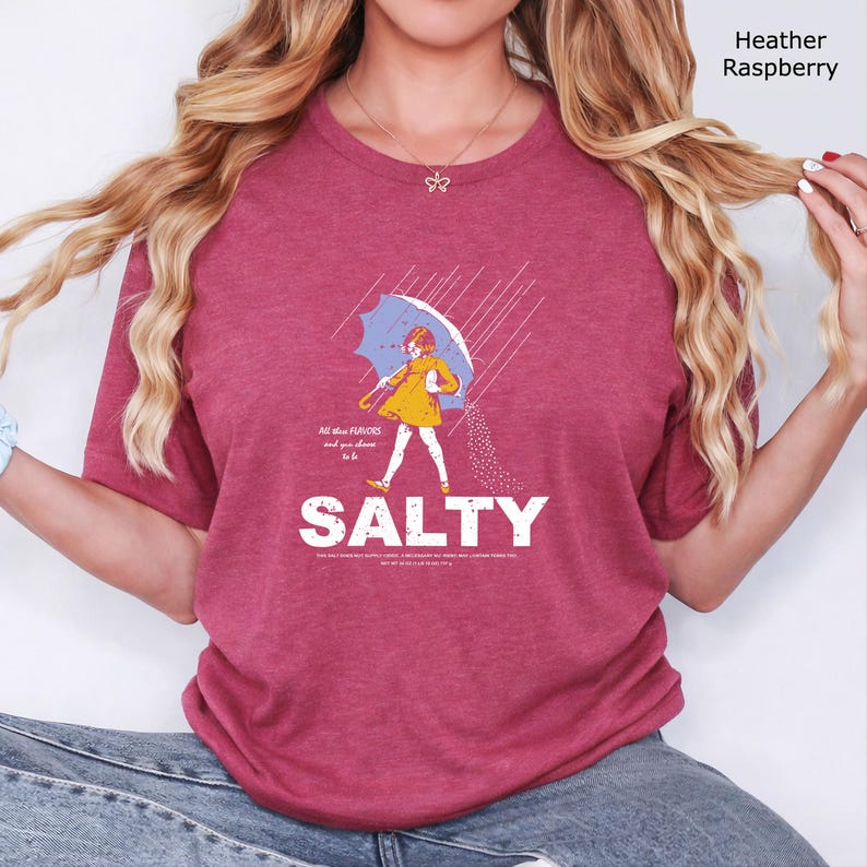 May include: A heather raspberry t-shirt featuring a graphic of a person in a yellow raincoat holding a blue umbrella in the rain, with the text "All these FLAVORS and you choose to be SALTY" and additional small text printed below the main word.