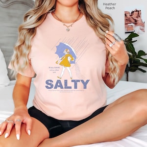 May include: A heather peach t-shirt featuring a graphic of a person in a yellow raincoat holding a blue umbrella, with the text SALTY printed in large blue letters below. The design includes rain lines and a scattering of salt grains.