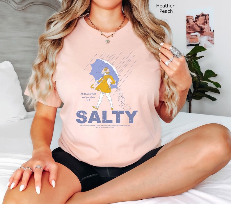 May include: A heather peach t-shirt featuring a graphic of a person in a yellow raincoat holding a blue umbrella, with the text SALTY printed in large blue letters below. The design includes rain lines and a scattering of salt grains.