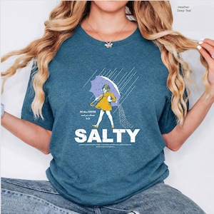 May include: A heather deep teal t-shirt featuring a graphic of a child in a yellow raincoat holding a purple umbrella in the rain. The text reads All these FLAVORS and you choose to be SALTY, with additional small text below the word SALTY.