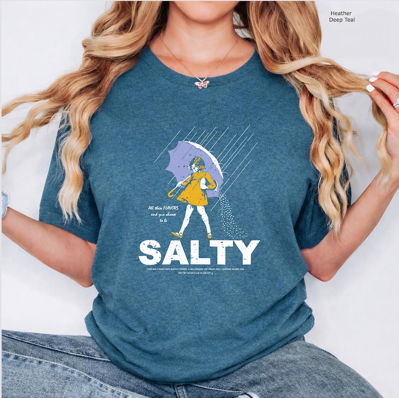 May include: A heather deep teal t-shirt featuring a graphic of a child in a yellow raincoat holding a purple umbrella in the rain. The text reads All these FLAVORS and you choose to be SALTY, with additional small text below the word SALTY.