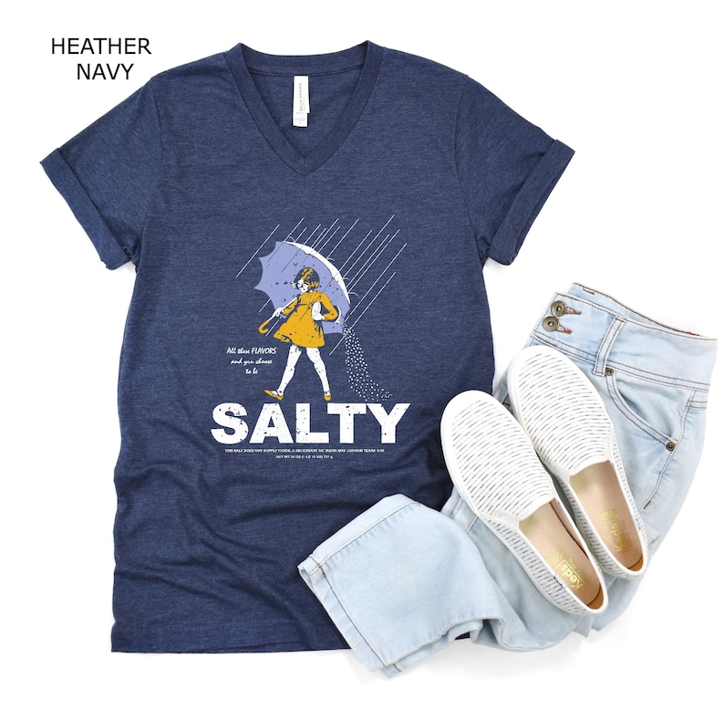 May include: A heather navy V-neck t-shirt featuring a graphic of a person in a yellow raincoat holding an umbrella in the rain, with the text "All the flavours and you choose to be SALTY" printed in white, paired with light blue jeans and white slip-on shoes.