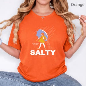 May include: An orange short-sleeve t-shirt featuring a graphic of a person in a yellow dress holding a blue umbrella in the rain. The graphic includes the text "All the flavours and you choose to be SALTY" with additional small text below.