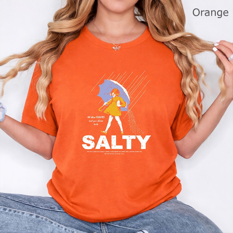 May include: An orange short-sleeve t-shirt featuring a graphic of a person in a yellow dress holding a blue umbrella in the rain. The graphic includes the text "All the flavours and you choose to be SALTY" with additional small text below.