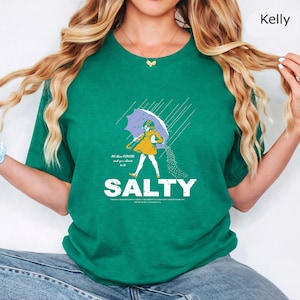 May include: A kelly green t-shirt featuring a graphic of a person in a yellow dress holding a purple umbrella in the rain, with salt pouring from the umbrella. The text reads "All these FLAVORS and you choose to be SALTY" in white lettering.