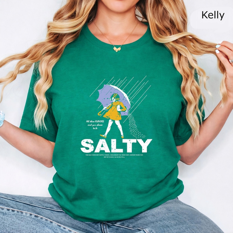 May include: A kelly green t-shirt featuring a graphic of a person in a yellow dress holding a purple umbrella in the rain, with salt pouring from the umbrella. The text reads "All these FLAVORS and you choose to be SALTY" in white lettering.