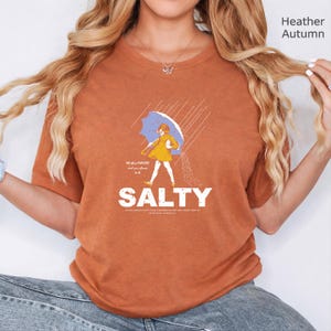 May include: A heather autumn orange t-shirt featuring a graphic of a person in a yellow raincoat holding a blue umbrella in the rain, with the text "All these FLAVORS and you choose to be SALTY" printed in white lettering on the front.