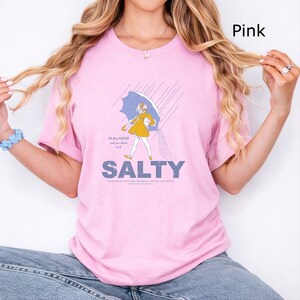 May include: A light pink t-shirt featuring a graphic of a person in a yellow raincoat holding a blue umbrella in the rain. The text reads "All these FLAVORS and you choose to be SALTY" with additional small text below the word SALTY.