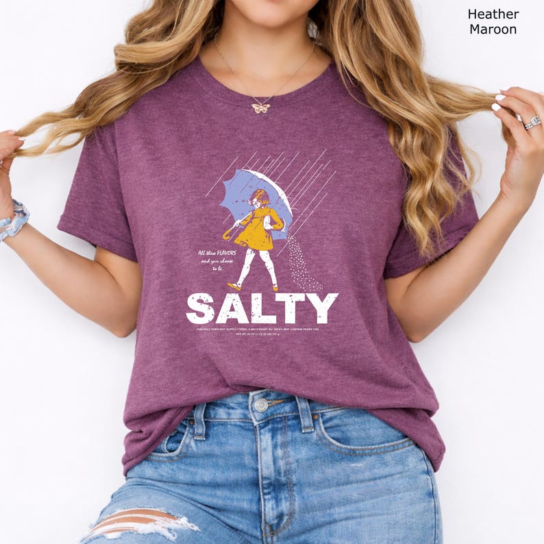 May include: A heather maroon short-sleeve t-shirt featuring a graphic of a person in a yellow raincoat holding an umbrella in the rain, with the text "All the flavours... and you choose to be SALTY" printed in white below the illustration.