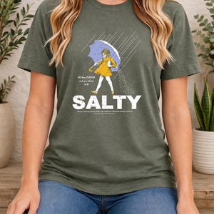 May include: An olive green short-sleeve t-shirt featuring a graphic of a child in a yellow raincoat holding a blue umbrella in the rain, pouring salt. The text reads "All these FLAVORS and you choose to be SALTY" with additional small print text.