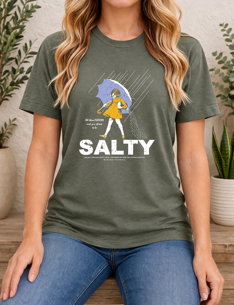 May include: An olive green short-sleeve t-shirt featuring a graphic of a child in a yellow raincoat holding a blue umbrella in the rain, pouring salt. The text reads "All these FLAVORS and you choose to be SALTY" with additional small print text.