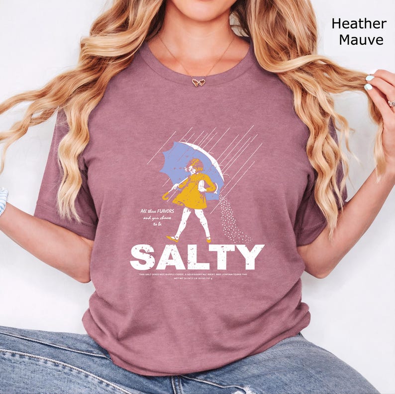 May include: A heather mauve t-shirt featuring a graphic of a person in a yellow raincoat holding a blue umbrella in the rain, pouring salt. The text reads All these FLAVORS and you choose to be SALTY, with smaller text below the word SALTY.