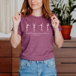 A woman wearing a purple t-shirt with a graphic design of four human figures in various poses, standing in front of a wooden dresser with a potted plant.
