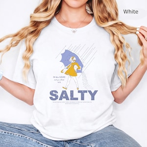 May include: A white t-shirt featuring a graphic of a person in a yellow raincoat holding a blue umbrella in the rain, with the text "All these FLAVORS and you choose to be SALTY" and "THIS SALT DOES NOT SUPPLY IODIDE, A NECESSARY NUTRIENT. NET WT 26 OZ (1 LB 10 OZ) 737 g."