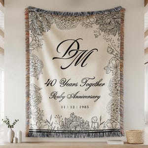 May include: A cream-colored woven throw blanket with black floral border and accents. The blanket features the initials "DM" and the text "40 Years Together Ruby Anniversary 11|12|1985". The blanket has a fringed edge.