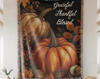 May include: A woven throw blanket featuring a fall-themed design with two pumpkins, leaves, and berries. The words "Grateful, Thankful, Blessed" are written in script. The blanket has a black background and a fringed edge.