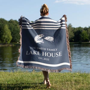 May include: A navy blue and white striped throw blanket with a fringed edge. The blanket features two white fish illustrations and the text "THE SMITH FAMILY LAKE HOUSE EST. 2025". The blanket is held up in front of a lake.