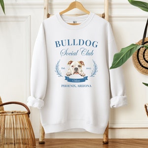 Puede incluir: Sudadera blanca con el texto "BULLDOG Social Club" en azul. El dise&ntilde;o incluye una ilustraci&oacute;n de bulldog, una corona de laurel y el texto "Chloe, Phoenix, Arizona". La sudadera est&aacute; colgada en una percha de madera.