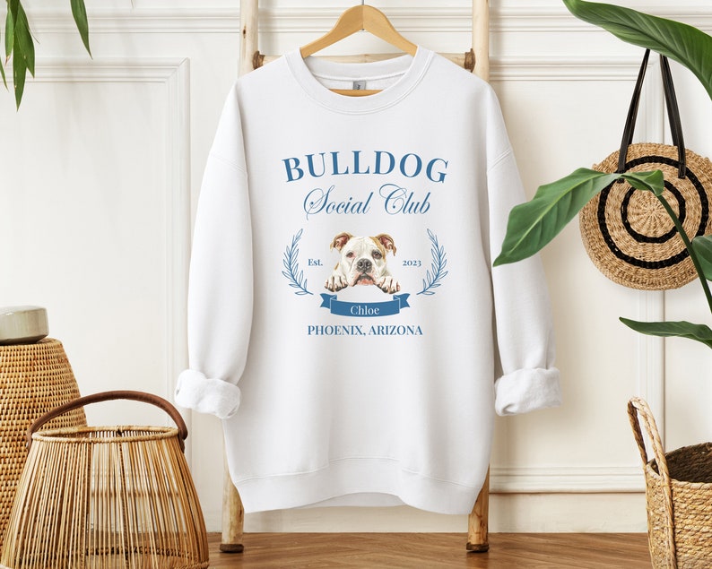 Puede incluir: Sudadera blanca con el texto "BULLDOG Social Club" en azul. El dise&ntilde;o incluye una ilustraci&oacute;n de bulldog, una corona de laurel y el texto "Chloe, Phoenix, Arizona". La sudadera est&aacute; colgada en una percha de madera.