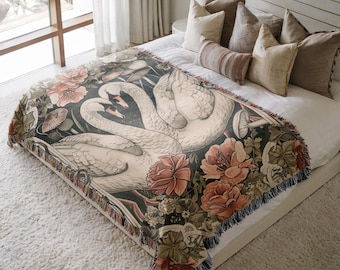 May include: A woven throw blanket featuring a design of two swans with intertwined necks, surrounded by flowers and foliage in shades of peach, green, and cream. The blanket has a dark background and a fringed edge, adding a decorative touch to the bedding.