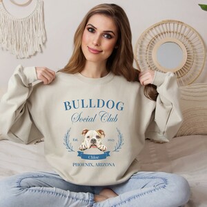 Puede incluir: Sudadera beige con el texto "BULLDOG Social Club" en azul y un gr&aacute;fico de bulldog. La sudadera tambi&eacute;n incluye el texto "Est. 2023" y "Chloe Phoenix, Arizona". La sudadera es de cuello redondo.