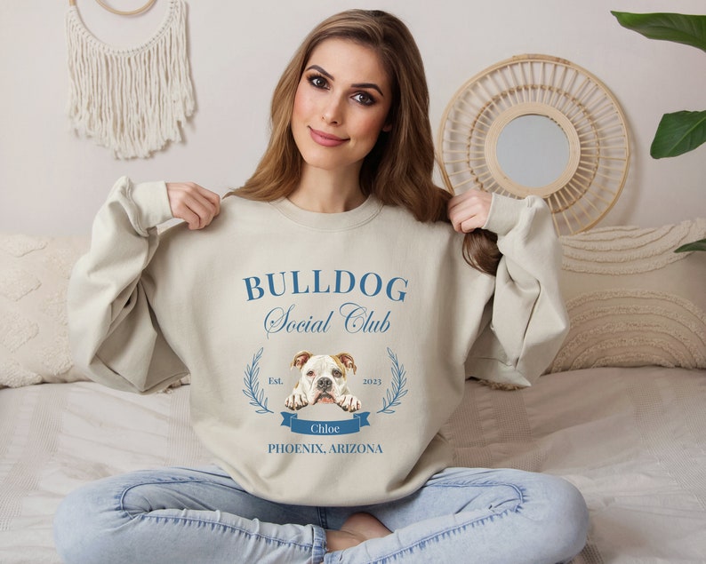 Puede incluir: Sudadera beige con el texto "BULLDOG Social Club" en azul y un gr&aacute;fico de bulldog. La sudadera tambi&eacute;n incluye el texto "Est. 2023" y "Chloe Phoenix, Arizona". La sudadera es de cuello redondo.