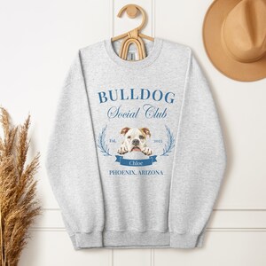 Puede incluir: Sudadera gris claro con el texto "BULLDOG Social Club" en azul. Debajo, un gr&aacute;fico de un bulldog y el nombre "Chloe". La sudadera est&aacute; colgada de una percha de madera.