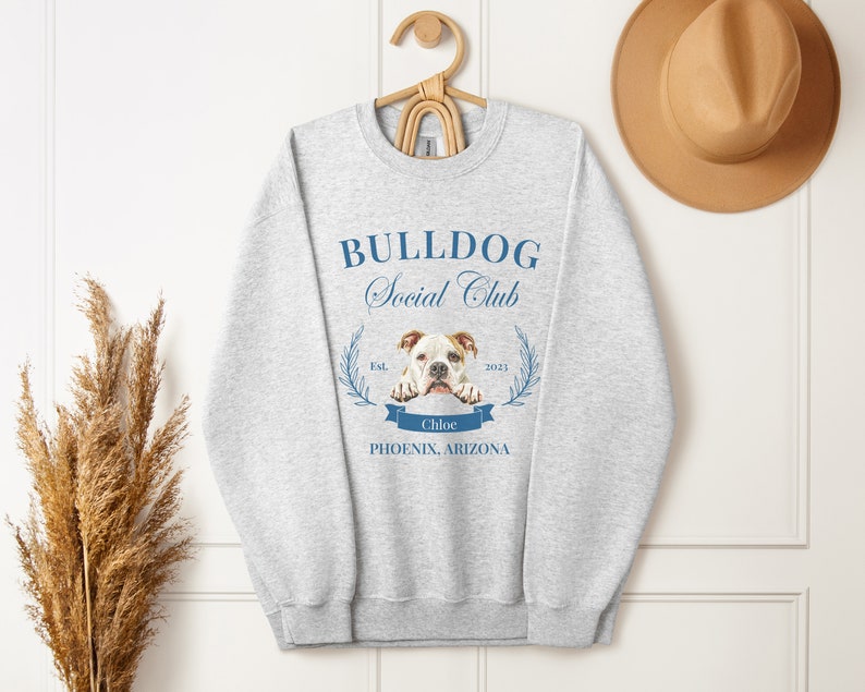 Puede incluir: Sudadera gris claro con el texto "BULLDOG Social Club" en azul. Debajo, un gr&aacute;fico de un bulldog y el nombre "Chloe". La sudadera est&aacute; colgada de una percha de madera.