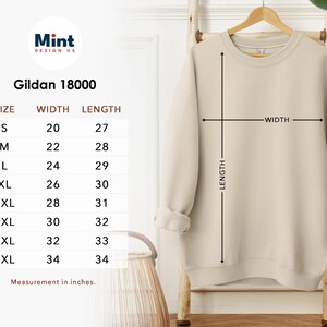 Puede incluir: Una sudadera beige Gildan 18000 colgada en un perchero de madera. La imagen incluye una tabla de tallas con medidas en cent&iacute;metros. El ancho de la sudadera var&iacute;a de 50,8 cm a 86,4 cm, y el largo de 68,6 cm a 86,4 cm. Las tallas van de la S a la 5XL.