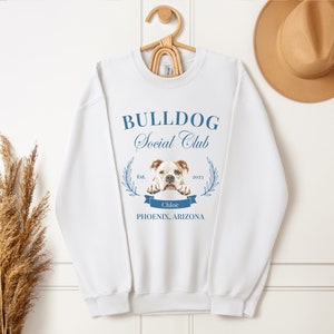 Puede incluir: Sudadera blanca con el texto azul "BULLDOG Social Club". El dise&ntilde;o incluye una imagen de bulldog, una pancarta con el nombre "Chloe" y la ubicaci&oacute;n "PHOENIX, ARIZONA". La sudadera est&aacute; colgada en una percha de madera.