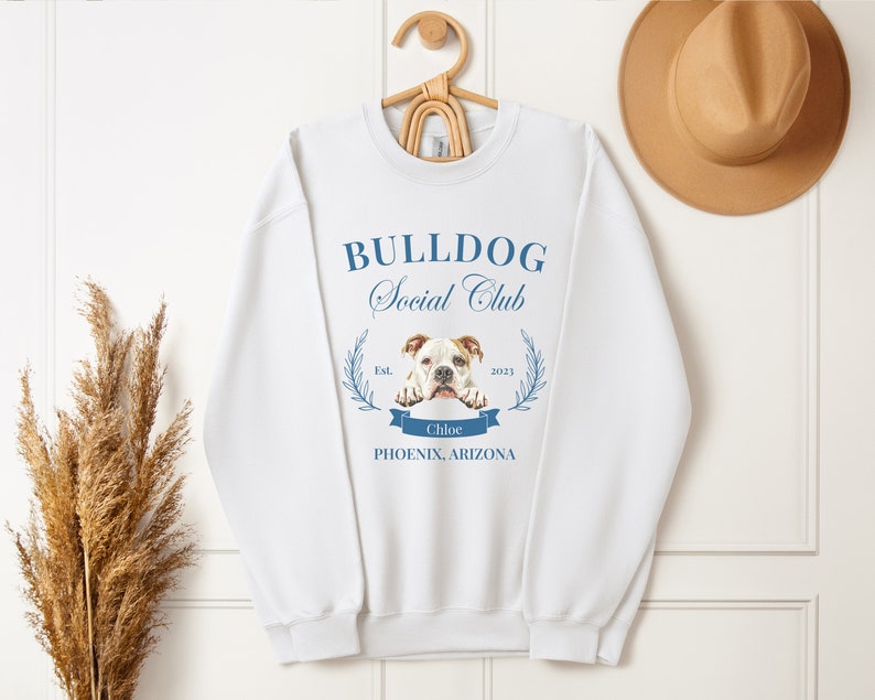 Puede incluir: Sudadera blanca con el texto azul "BULLDOG Social Club". El dise&ntilde;o incluye una imagen de bulldog, una pancarta con el nombre "Chloe" y la ubicaci&oacute;n "PHOENIX, ARIZONA". La sudadera est&aacute; colgada en una percha de madera.