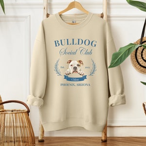 Puede incluir: Sudadera color crema con el texto azul "BULLDOG Social Club". El dise&ntilde;o incluye una ilustraci&oacute;n de bulldog, una pancarta con el nombre "Chloe" y la ubicaci&oacute;n "Phoenix, Arizona". La sudadera est&aacute; colgada en un perchero de madera.