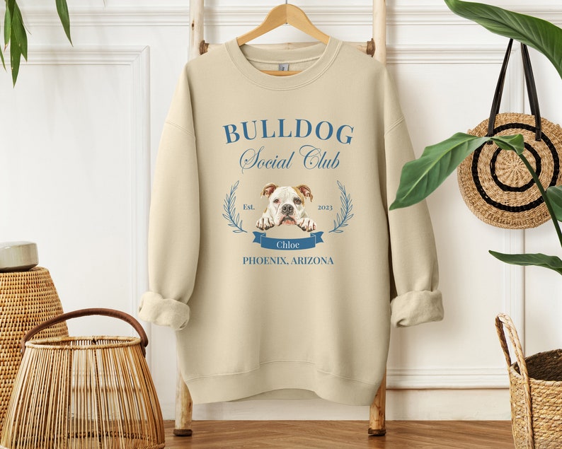 Puede incluir: Sudadera color crema con el texto azul "BULLDOG Social Club". El dise&ntilde;o incluye una ilustraci&oacute;n de bulldog, una pancarta con el nombre "Chloe" y la ubicaci&oacute;n "Phoenix, Arizona". La sudadera est&aacute; colgada en un perchero de madera.