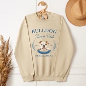 Puede incluir: Sudadera beige con el texto "BULLDOG Social Club" en escritura azul. Una imagen de bulldog est&aacute; centrada debajo del texto, con el nombre "Chloe" en una pancarta azul. La sudadera tambi&eacute;n incluye "PHOENIX, ARIZONA" y el a&ntilde;o "2023".