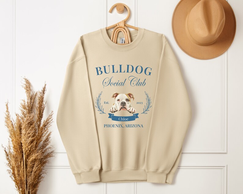 Puede incluir: Sudadera beige con el texto "BULLDOG Social Club" en escritura azul. Una imagen de bulldog est&aacute; centrada debajo del texto, con el nombre "Chloe" en una pancarta azul. La sudadera tambi&eacute;n incluye "PHOENIX, ARIZONA" y el a&ntilde;o "2023".